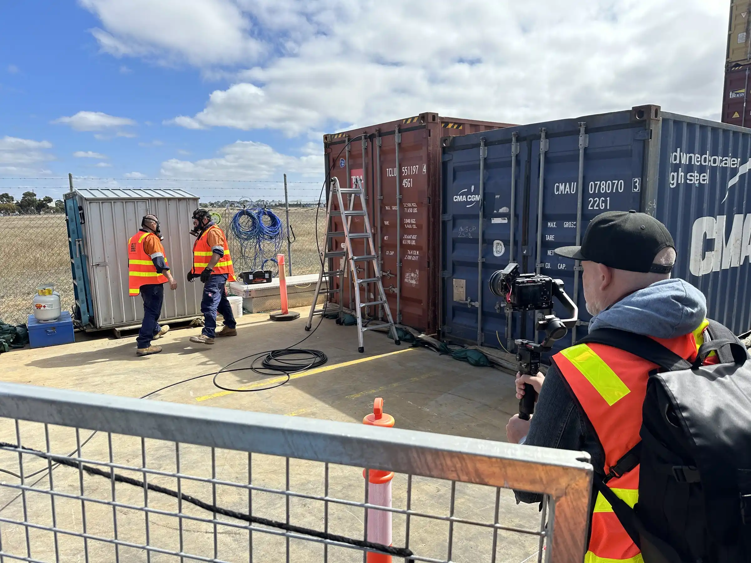 A HAUL videographer filming two depot workers with a gimbal camera between shipping containers